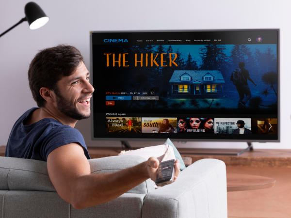 Person sitting on a sofa in a living room, holding a remote control pointed at a Smart TV showing a streaming application interface on the screen.