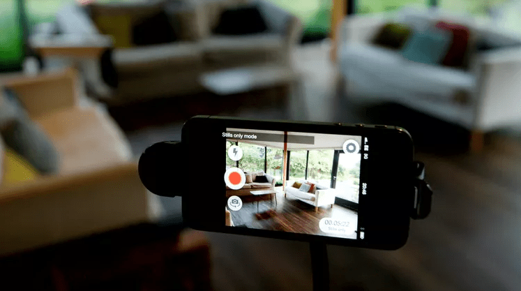 Smartphone on a stand running a security camera app and viewing a bright living room with sofas and large windows