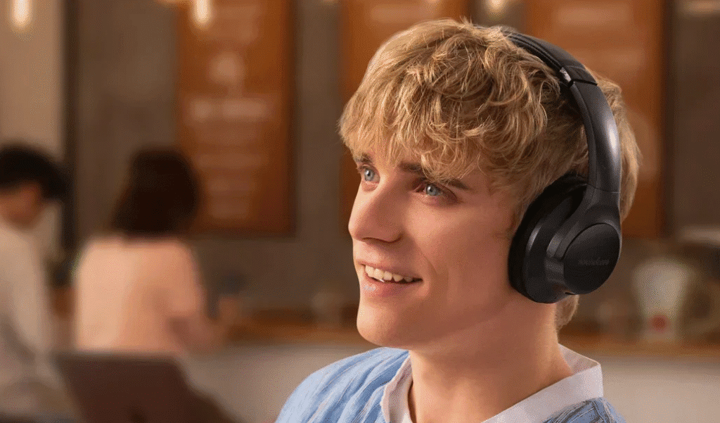 Smiling person wearing black over ear Bluetooth headphones in a cafe, looking slightly upward with a blurred background of other customers.