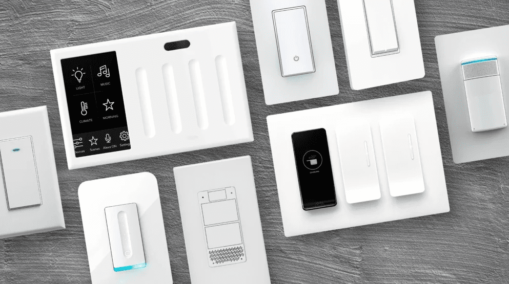 Overhead view of smart lighting controls on a tabletop, including wall switches, dimmer panels, a touch control screen, and sensor style devices.