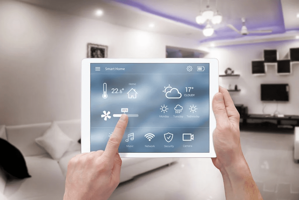 Hands holding a tablet showing a smart home dashboard, with a finger adjusting a lighting dimmer slider in a modern living room.