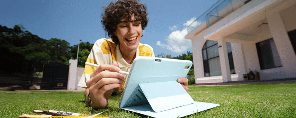 Person lying on a grassy lawn using a tablet in a stand case and writing with a stylus near a modern house.